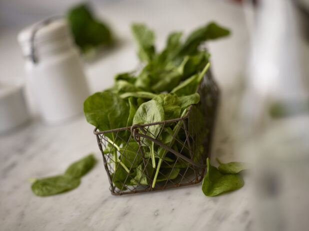 Whole fresh spinach in a decorative wire basket on a white marble table with white accessories in the background