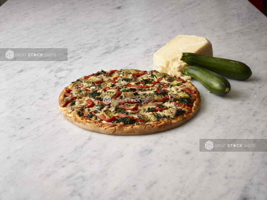 Whole pizza with zucchini, roasted red peppers and spinach with a block of parmesan and whole zucchini in the background on a white marble background