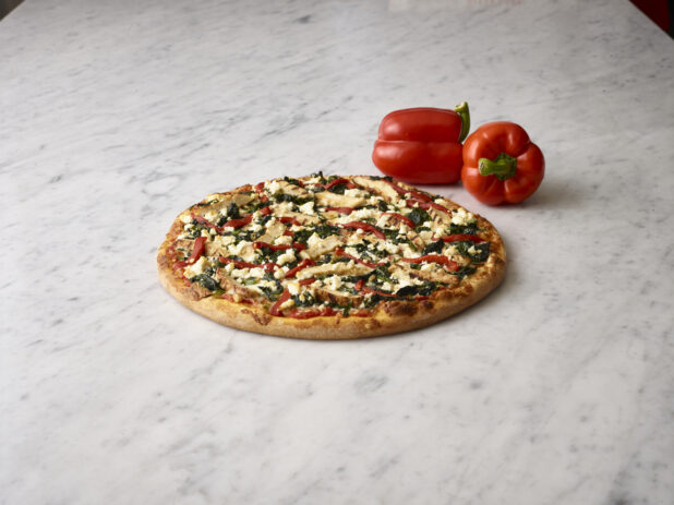 Whole pizza with chicken, spinach, roasted red peppers and feta cheese with whole fresh red bell peppers in the background all on a white marble background
