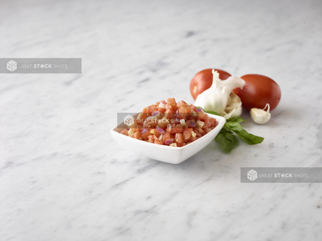 Close up view of bruschetta in a small white bowl with fresh basil, whole fresh garlic and whole fresh roma tomatoes behind