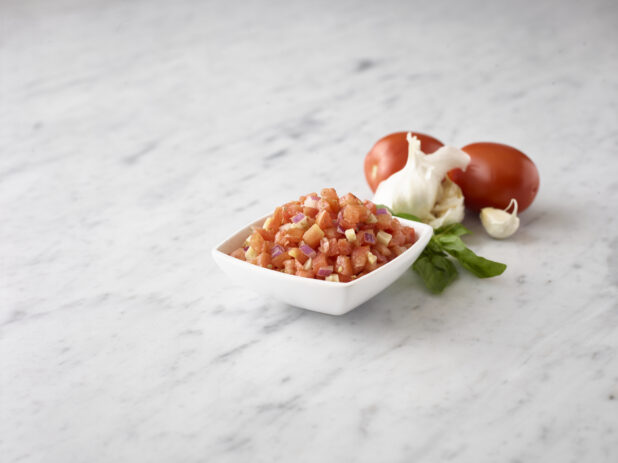 Close up view of bruschetta in a small white bowl with fresh basil, whole fresh garlic and whole fresh roma tomatoes behind