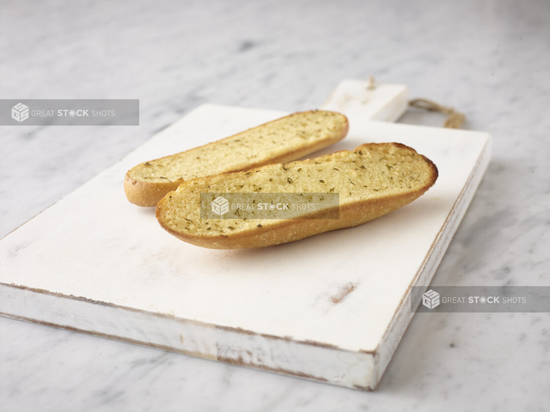 Whole garlic bread sliced in half on a white wooden board on a white marble background