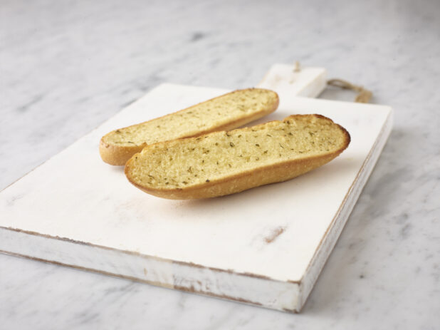 Whole garlic bread sliced in half on a white wooden board on a white marble background