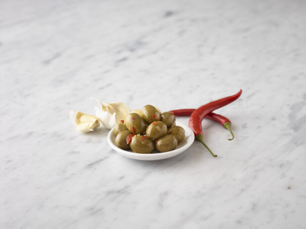 Whole green olives on a small white round plate with whole red chili peppers and garlic cloves on the sides all on a white marble background