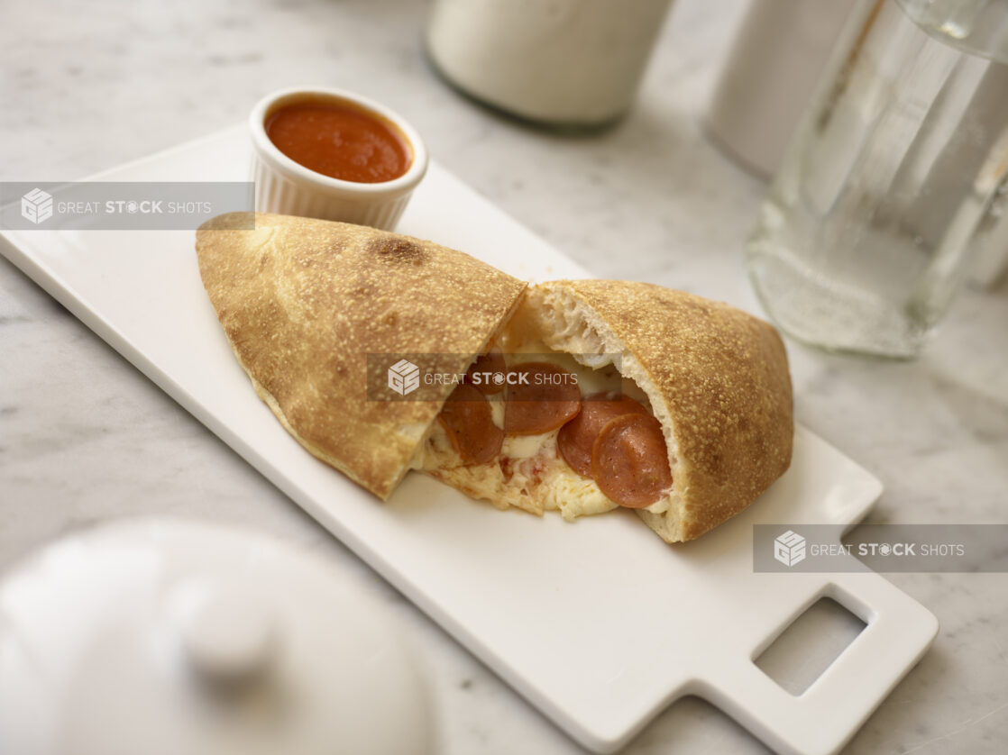 Pepperoni calzone on a white cutting board with a ramekin of tomato sauce on the side on a white marble background