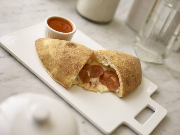 Pepperoni calzone on a white cutting board with a ramekin of tomato sauce on the side on a white marble background