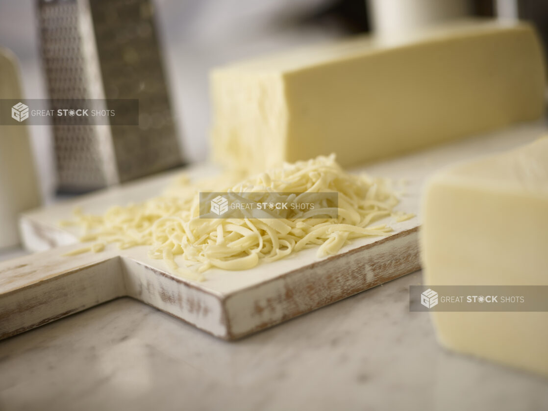 Whole brick of mozzarella cheese on a white wooden cutting board with shredded cheese in the foreground and a grater in the background