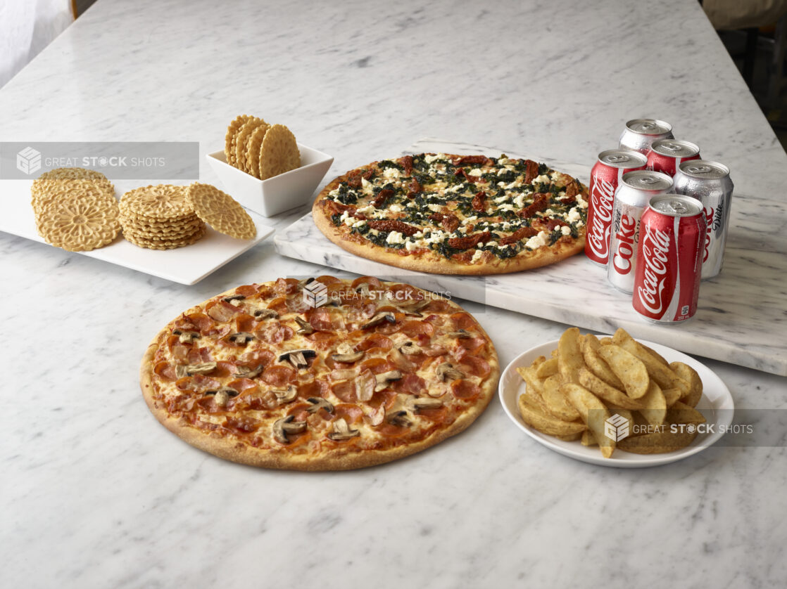 A whole pepperoni and mushroom pizza and a whole feta, sundried tomato and spinach pizza, surrounded by coke & diet cans, pizzelle and wedge french fries with a white marble background