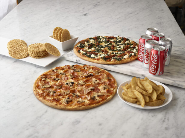 A whole pepperoni and mushroom pizza and a whole feta, sundried tomato and spinach pizza, surrounded by coke & diet cans, pizzelle and wedge french fries with a white marble background