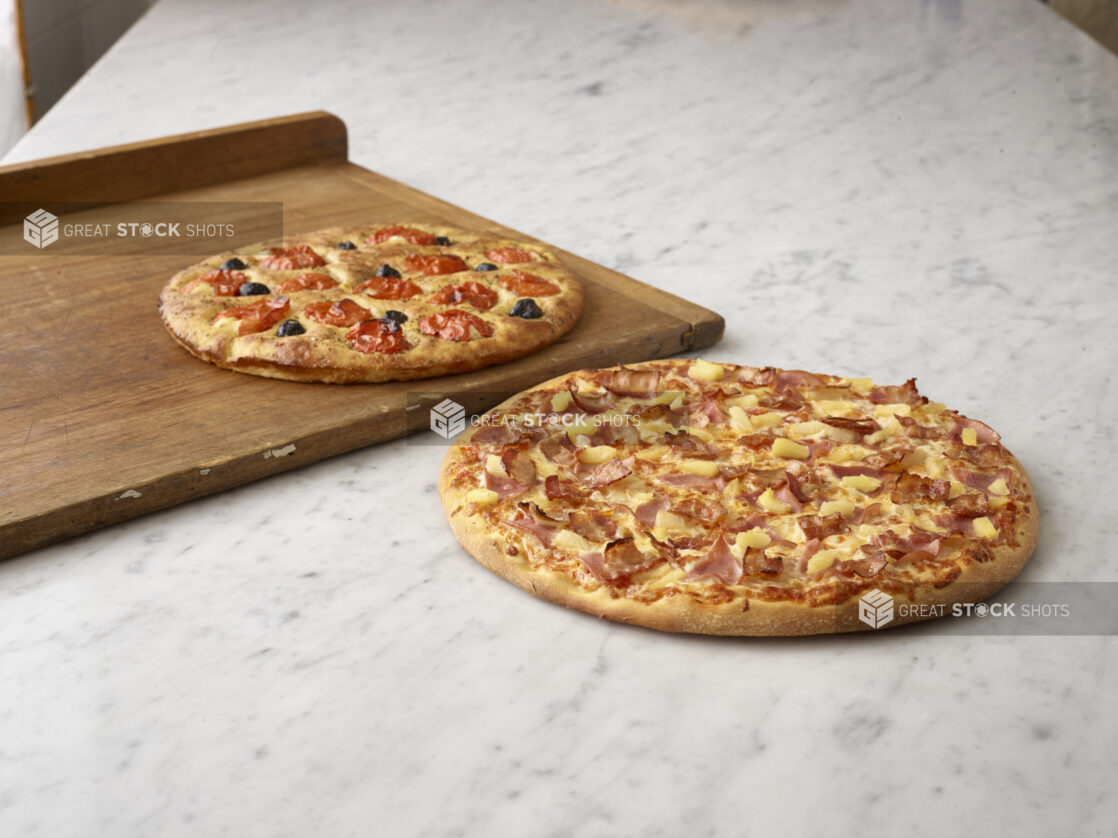 Whole Hawaiian pizza in the foreground on white marble and a tomato and olive round focaccia in the background on a wooden board