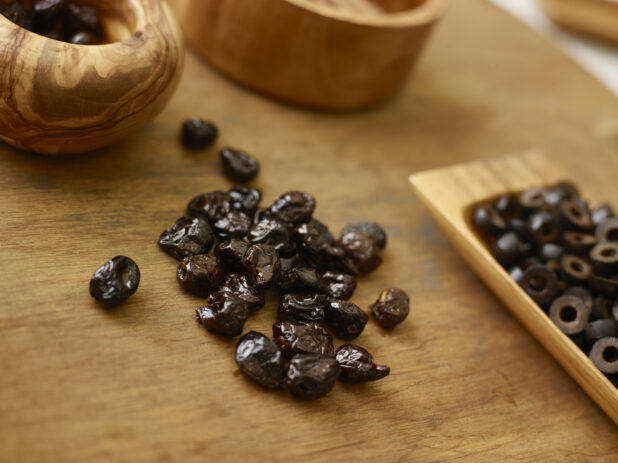Dried black olives beside a wooden tray of sliced black olives on a warm coloured wood surface