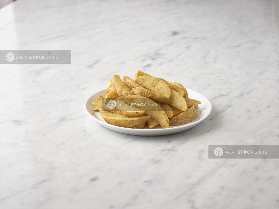 Mound of seasoned potato wedges on a round white plate on a white marble background