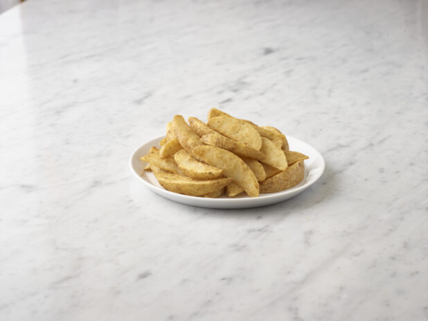 Mound of seasoned potato wedges on a round white plate on a white marble background