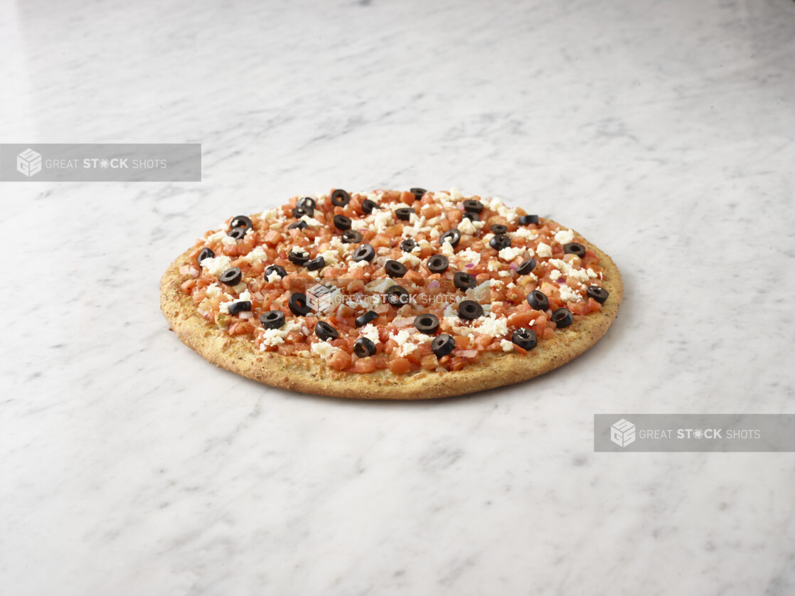 Whole unsliced chopped tomato, black olive, and feta pizza with sprig of basil alongside on a white marble background