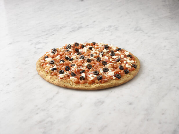 Whole unsliced chopped tomato, black olive, and feta pizza with sprig of basil alongside on a white marble background