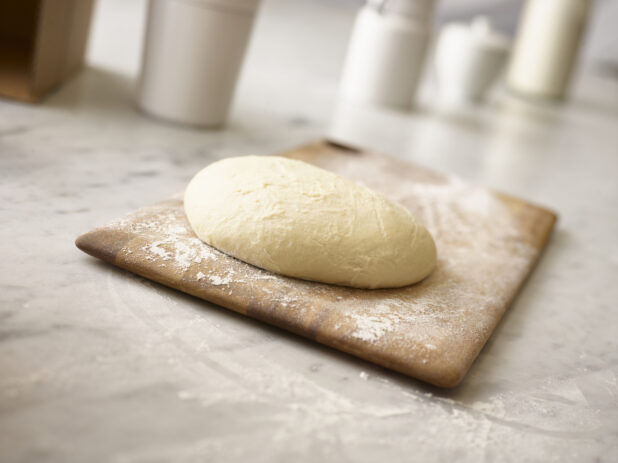 Pizza dough on a wooden cutting board on a marble table top shot in the foreground