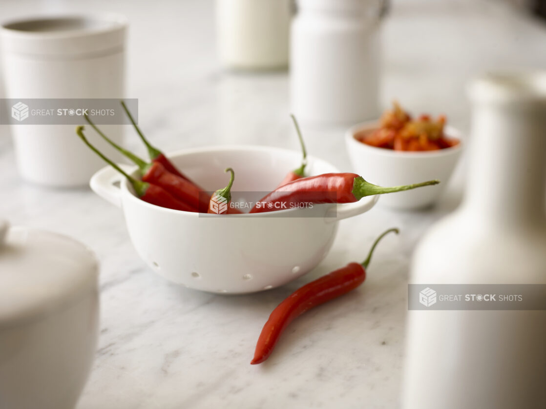 Whole fresh red chilli peppers in a white miniature ceramic sieve, white dishes and white marble background, close-up