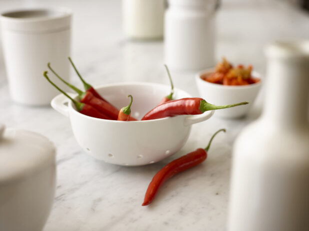 Whole fresh red chilli peppers in a white miniature ceramic sieve, white dishes and white marble background, close-up