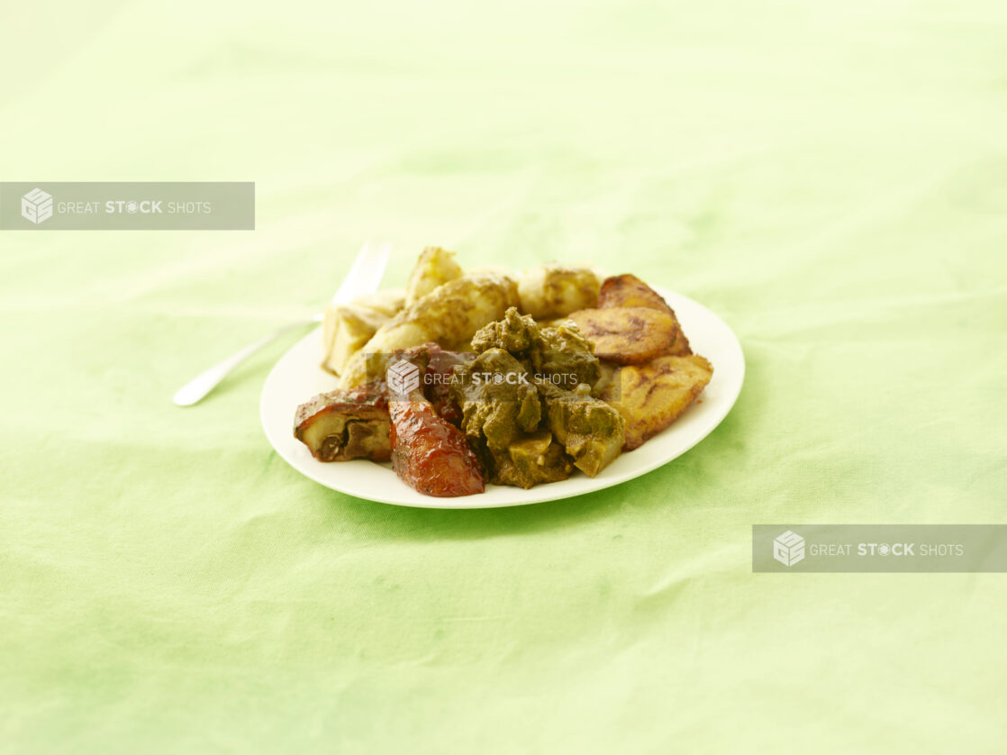 Curried goat and bbq chicken with fried plantain on a round white plate on a lemon/lime green tablecloth