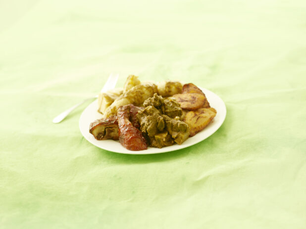 Curried goat and bbq chicken with fried plantain on a round white plate on a lemon/lime green tablecloth