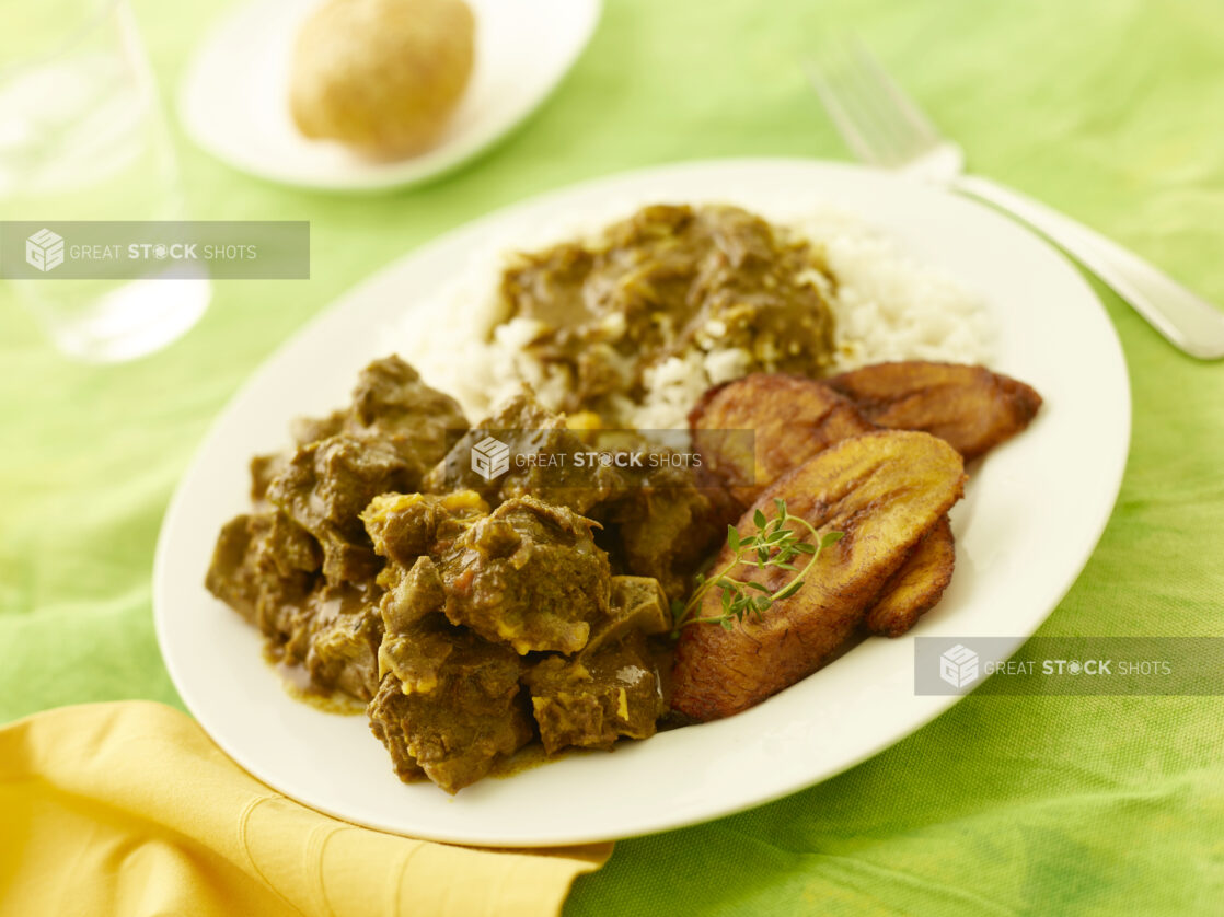 Caribbean curried goat with fried plantain and rice on a white plate on a lemon/lime green tablecloth