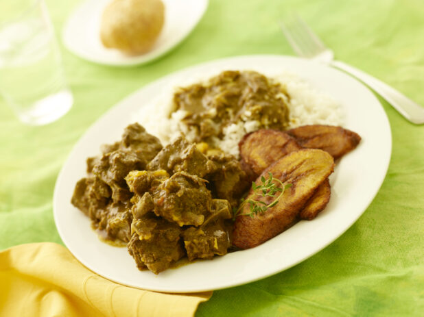 Caribbean curried goat with fried plantain and rice on a white plate on a lemon/lime green tablecloth