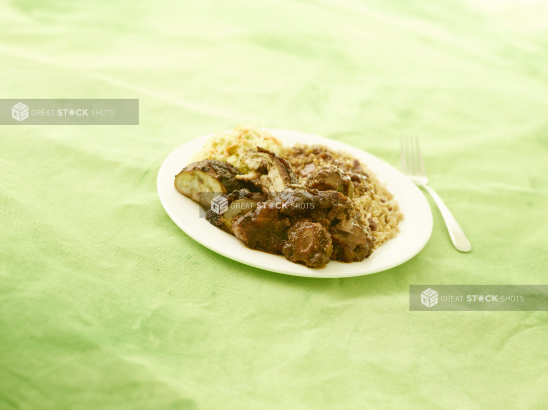 Caribbean jerk chicken, rice and peas and coleslaw on round white plate on lemon/lime green tablecloth