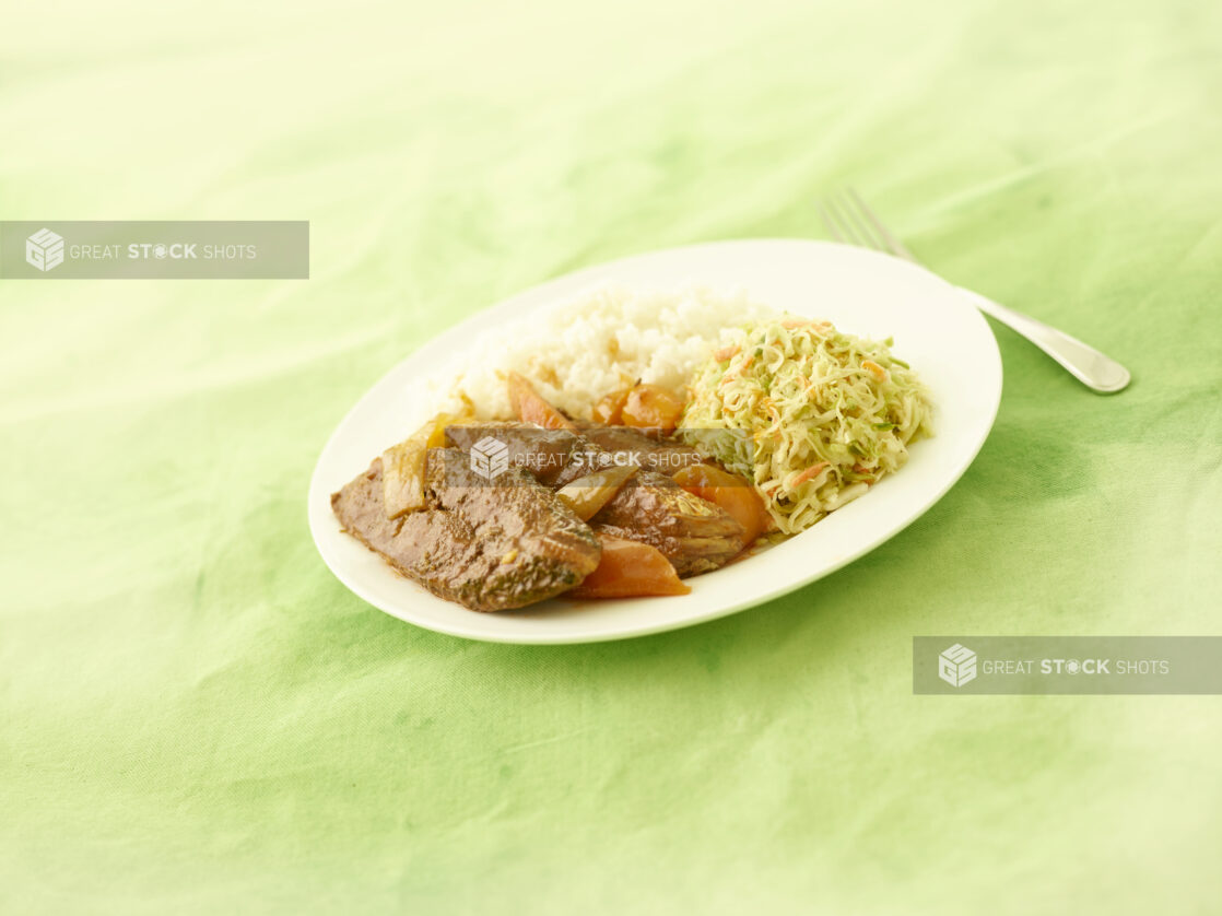 Caribbean brown stew fish with coleslaw and rice on an oval white plate on a lemon/lime green tablecloth