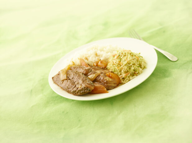 Caribbean brown stew fish with coleslaw and rice on an oval white plate on a lemon/lime green tablecloth