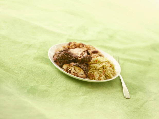 Caribbean jerk chicken with coleslaw and rice and peas on an oval white plate on a lemon/lime green background