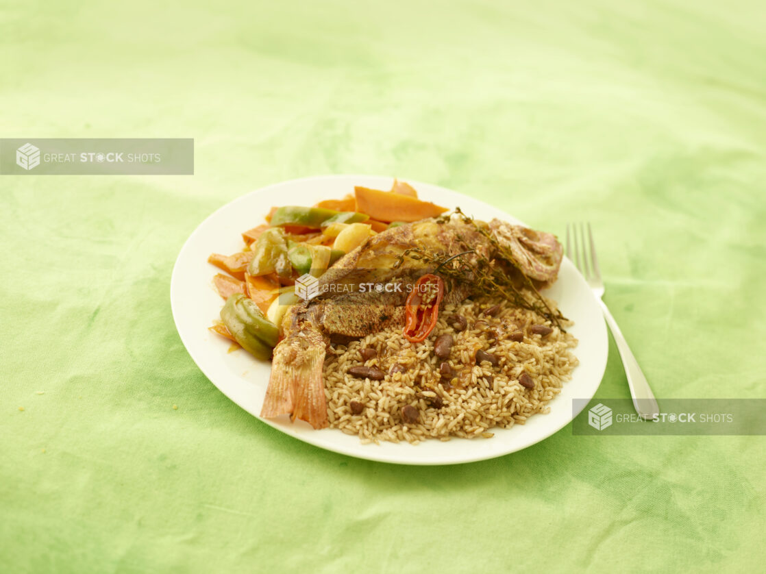 Caribbean fried fish with steamed veggies and rice and peas on a white plate with a lemon/lime green background