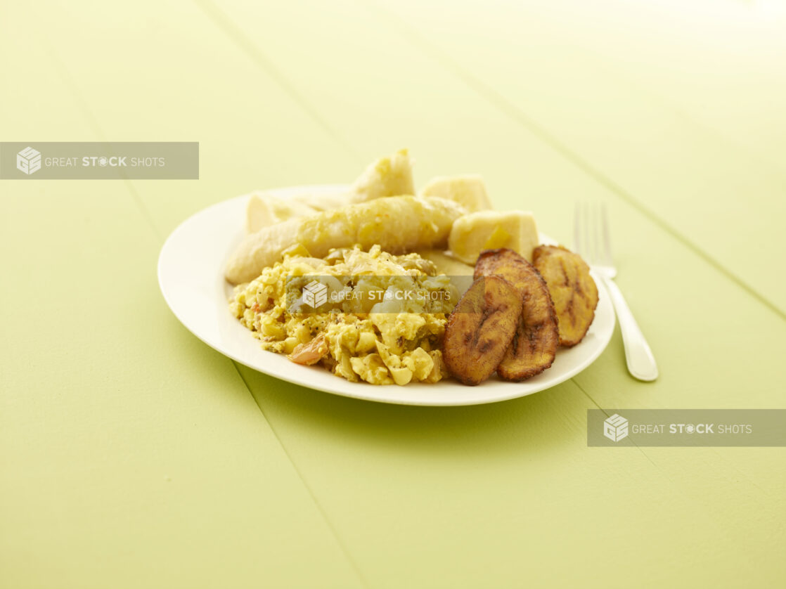 Caribbean ackee and salt fish with fried plantain, boiled banana, boiled dumpling on a white plate on a pale green table