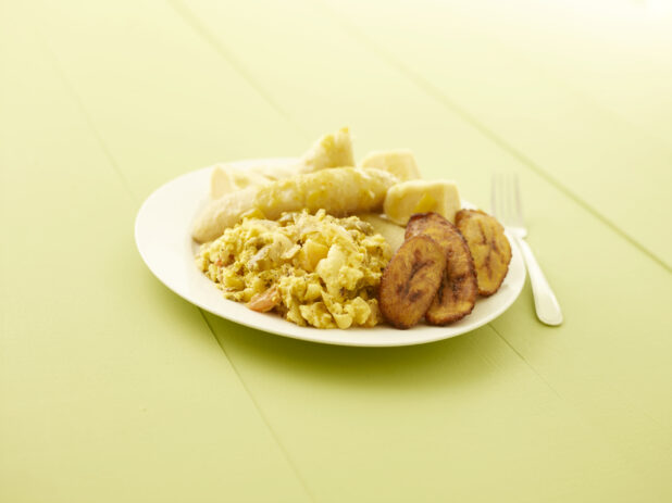 Caribbean ackee and salt fish with fried plantain, boiled banana, boiled dumpling on a white plate on a pale green table