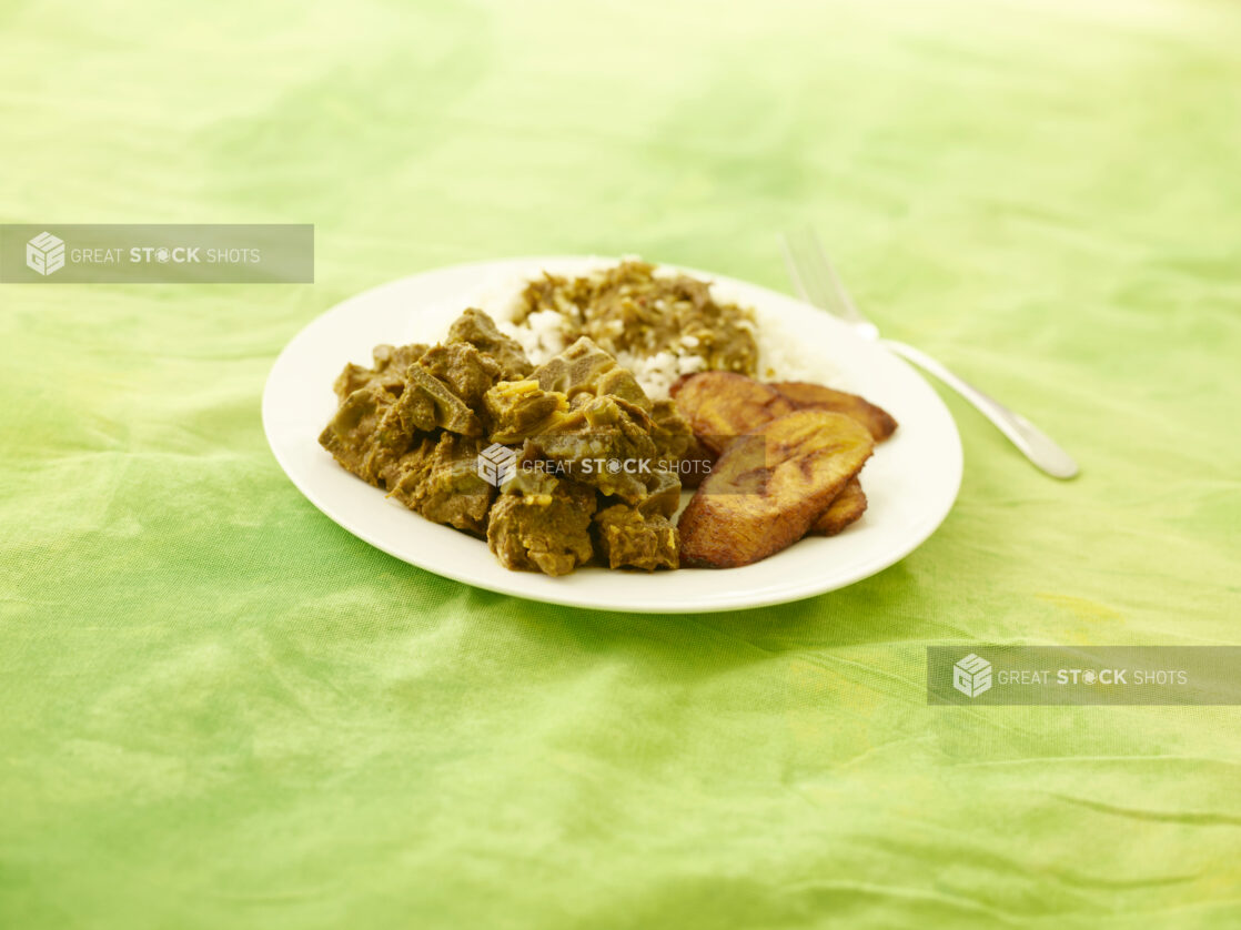 Caribbean curried goat with fried plantain and rice on a round white plate on a lemon/lime green tablecloth