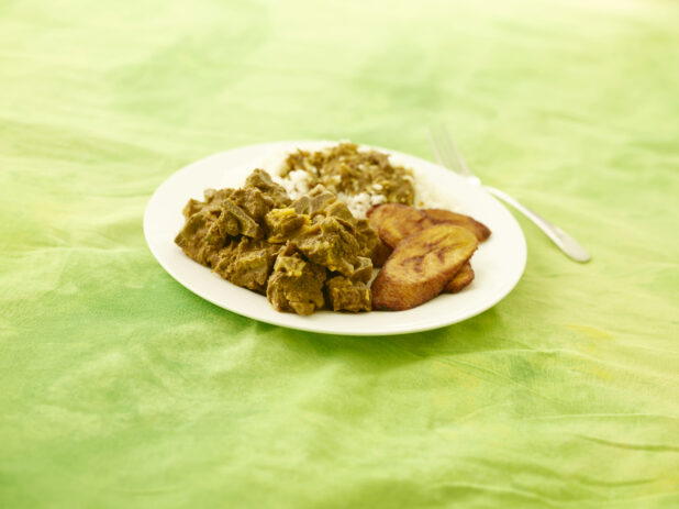 Caribbean curried goat with fried plantain and rice on a round white plate on a lemon/lime green tablecloth