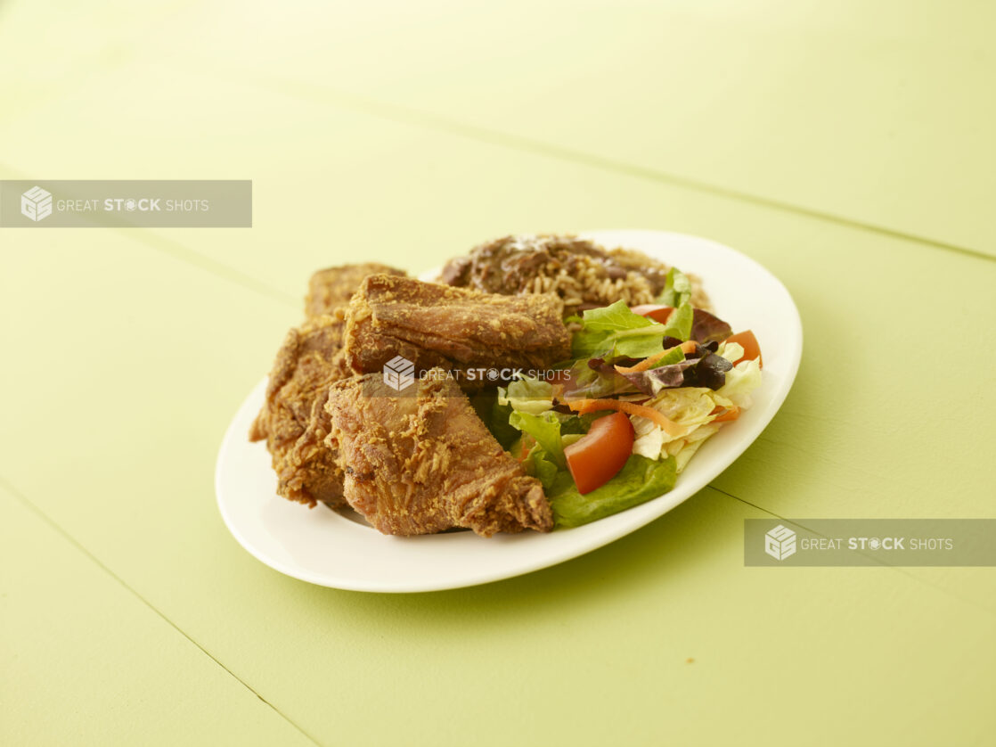 Caribbean fried chicken dinner with side salad and rice and peas on a white oval plate on a light green wooden table