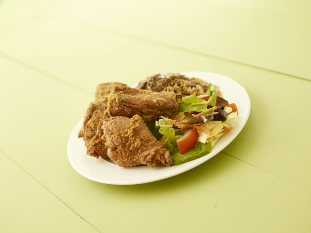 Caribbean fried chicken dinner with side salad and rice and peas on a white oval plate on a light green wooden table