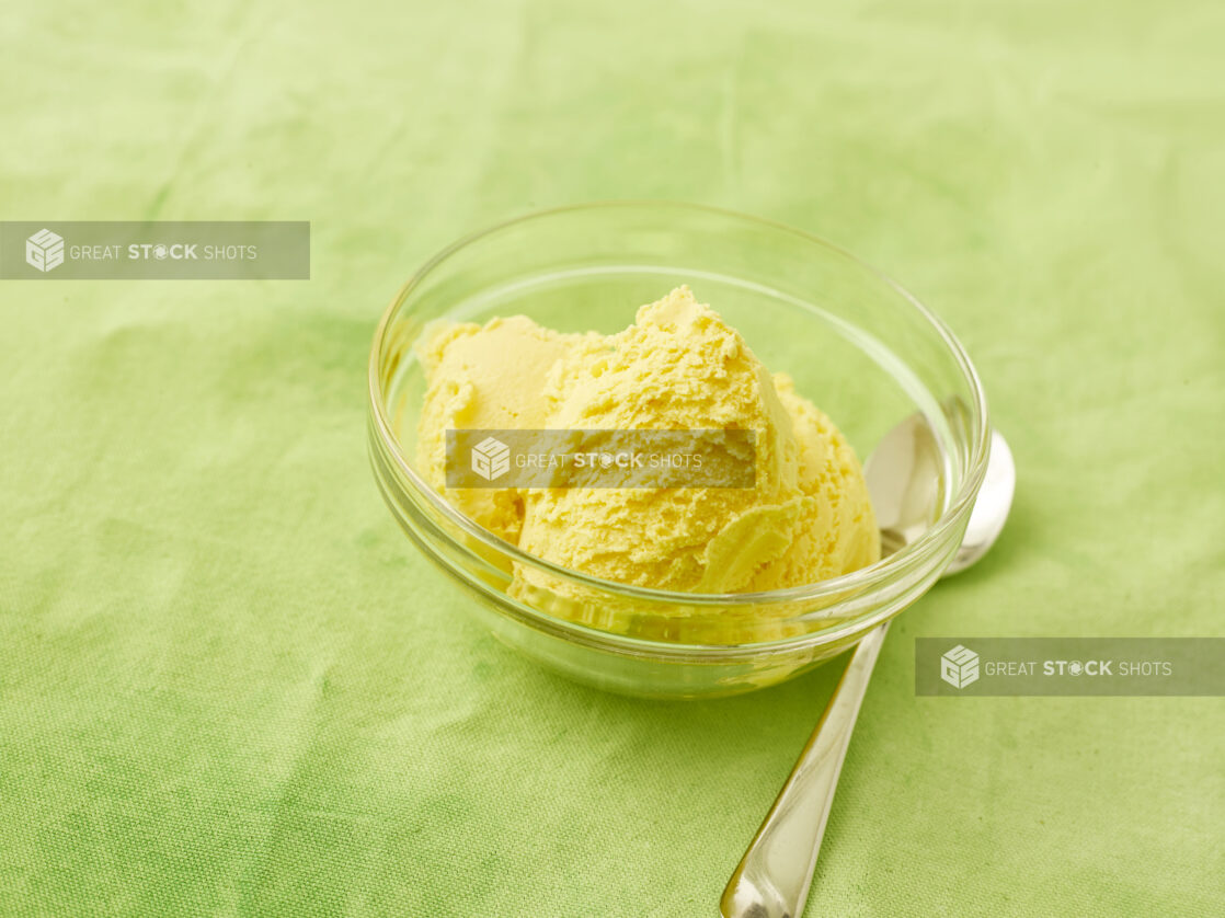 Mango ice cream in a small glass bowl with a dessert spoon on a lemon/lime green background