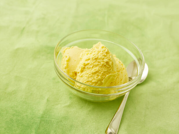 Mango ice cream in a small glass bowl with a dessert spoon on a lemon/lime green background