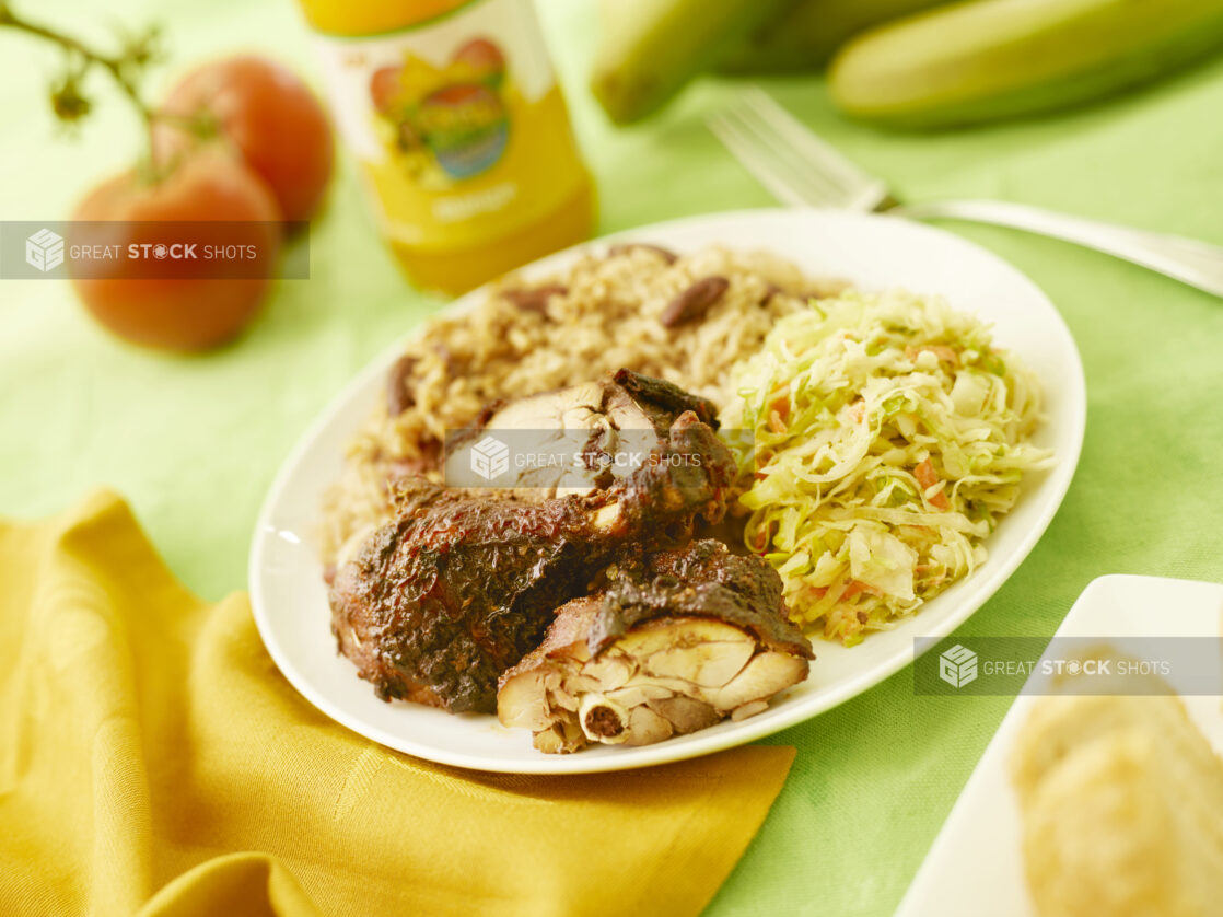 Jerk chicken dinner on a white round plate with fresh tomatoes, plantain and a beverage in the background on a lemon/lime green tablecloth