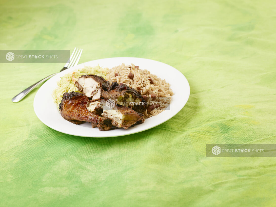 Caribbean jerk chicken dinner with rice and peas and coleslaw on a white round plate with a fork on a lemon/lime green tablecloth