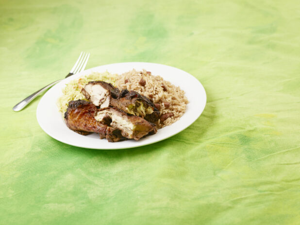 Caribbean jerk chicken dinner with rice and peas and coleslaw on a white round plate with a fork on a lemon/lime green tablecloth