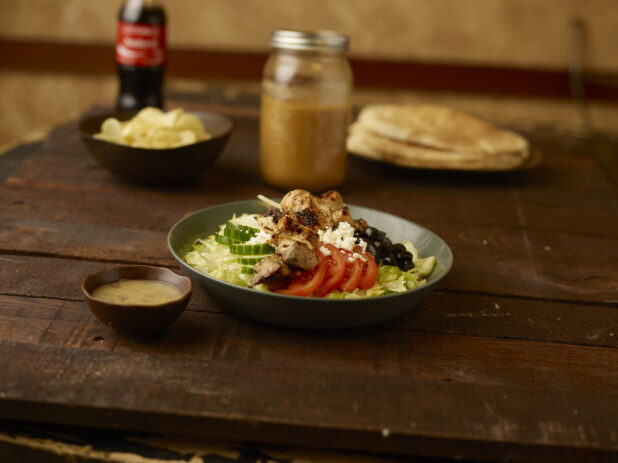 Greek salad with chicken on an aged wooden table, with small bowl of dressing in foreground with coke in bottle, potato chips in a side bowl and flatbread