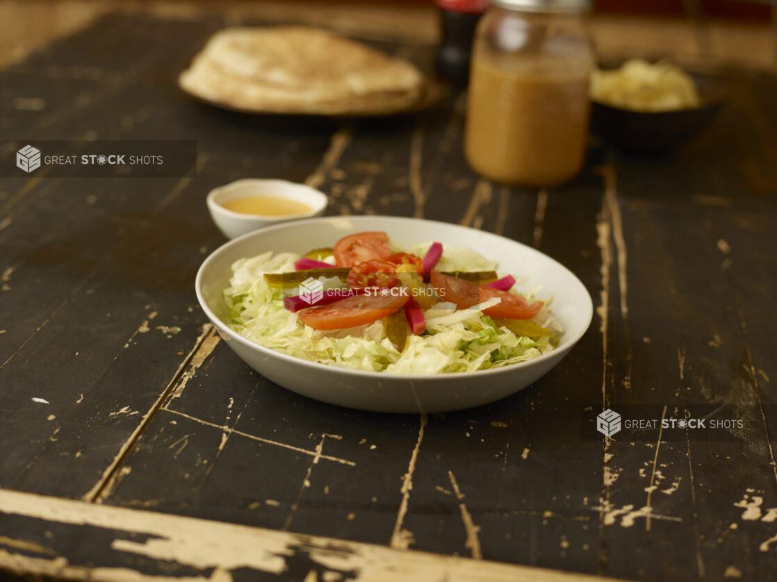 Side salad in the foreground with dressing on an aged wooden table with coke, flatbread and potato chips in the background