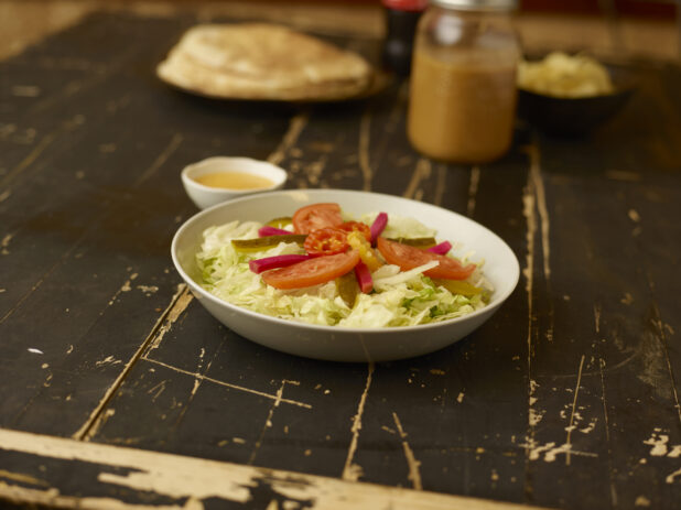 Side salad in the foreground with dressing on an aged wooden table with coke, flatbread and potato chips in the background