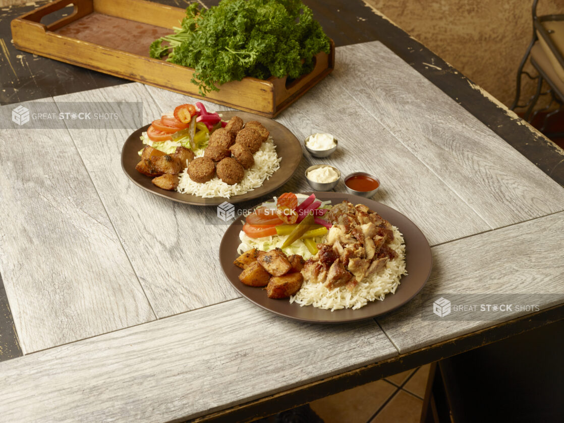 Chicken shawarma dinner and falafel dinners on round plates on a wooden table with fresh parsley in a wooden crate in the background