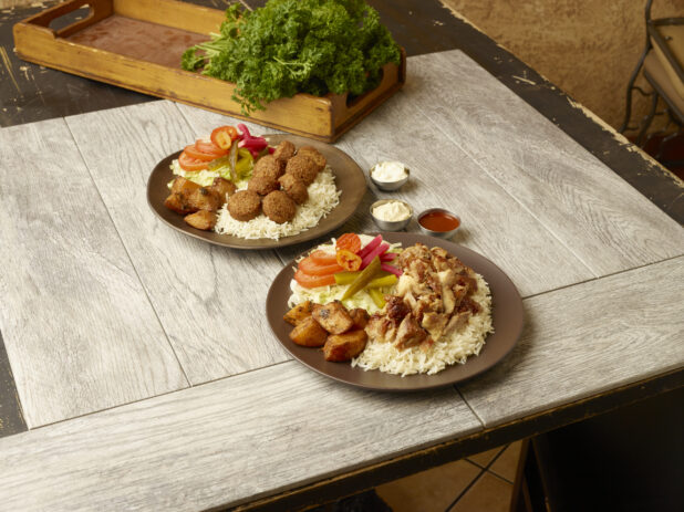 Chicken shawarma dinner and falafel dinners on round plates on a wooden table with fresh parsley in a wooden crate in the background
