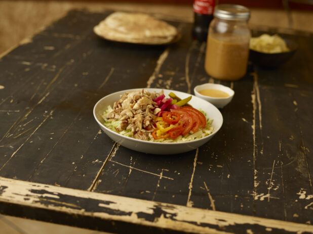 Chicken shawarma salad in the foreground, pitas, bowl of chips and a Coke bottle in the background all on a weathered table