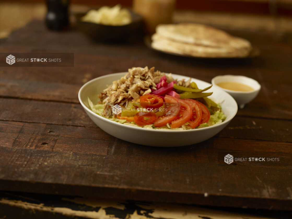 Chicken shawarma salad in the foreground with pitas, bowl of chips and dressing in the background all on a rustic wooden table