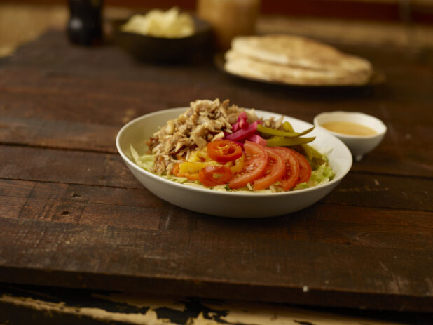 Chicken shawarma salad in the foreground with pitas, bowl of chips and dressing in the background all on a rustic wooden table